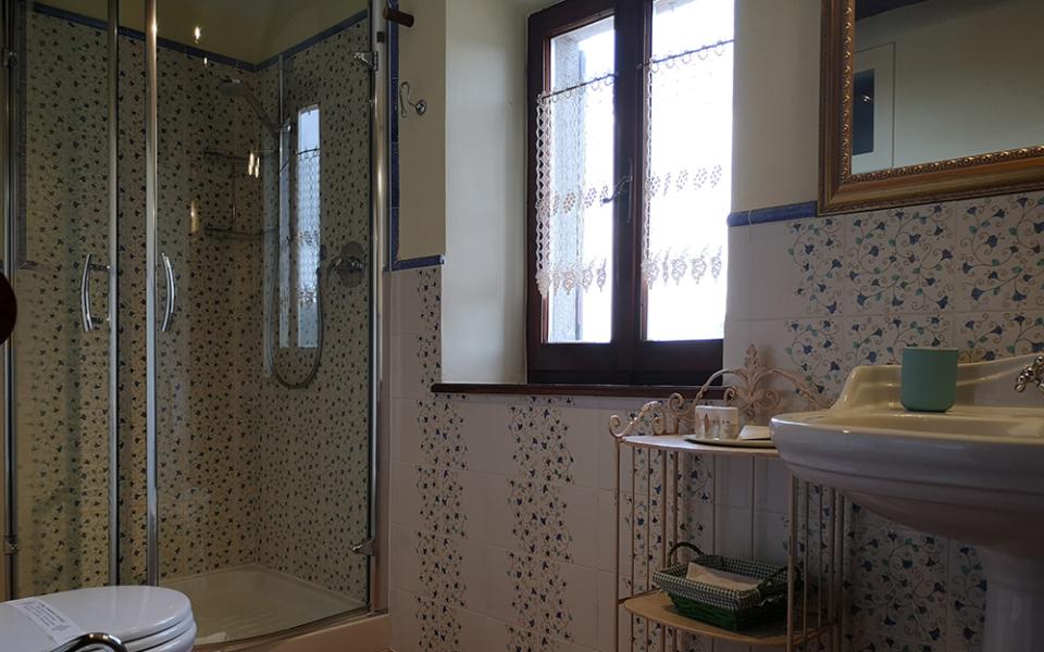 Bagno con doccia, piastrelle floreali, finestra, lavandino e scaffale con accessori.