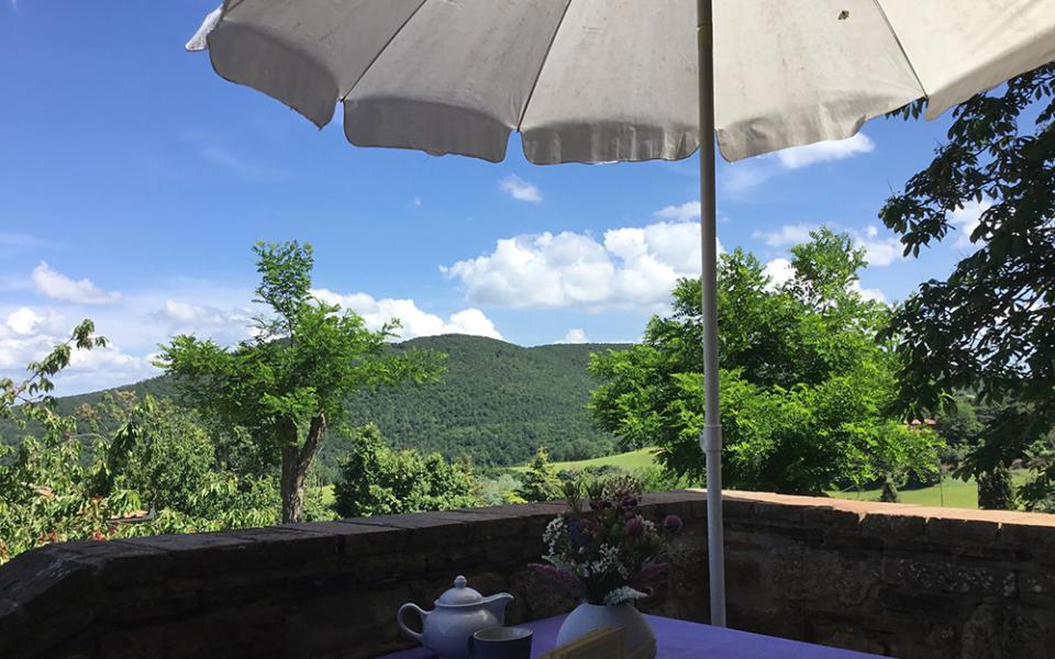 Terrazza con vista collinare, tavolo viola, tè e libro aperto sotto l'ombrellone.
