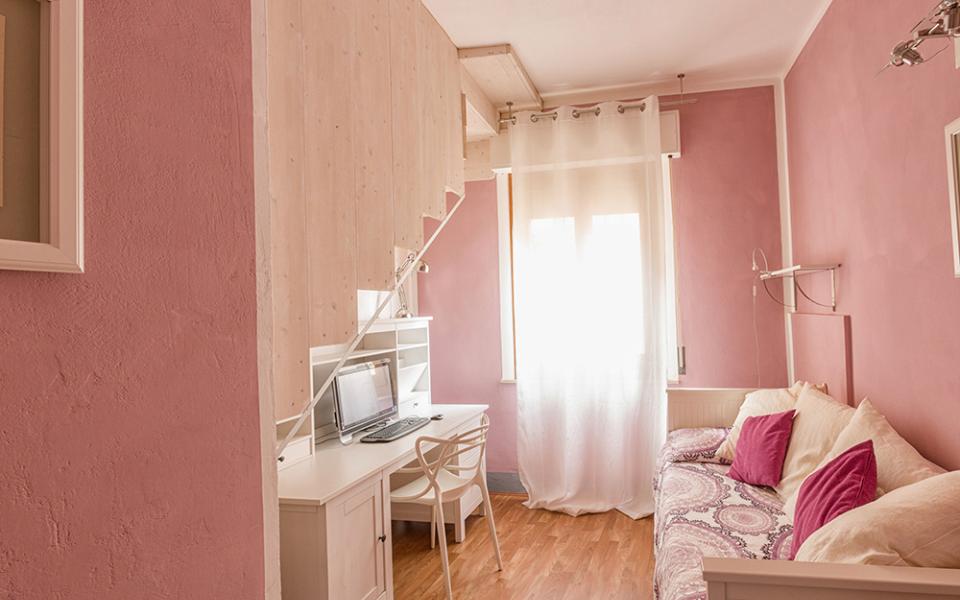Camera rosa con scrivania, divano e cuscini colorati, decorata con tende bianche.
