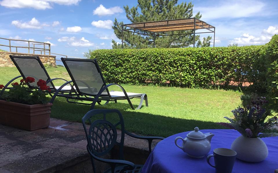 Giardino con sdraio, tavolo apparecchiato per il tè e fiori sotto il cielo azzurro.